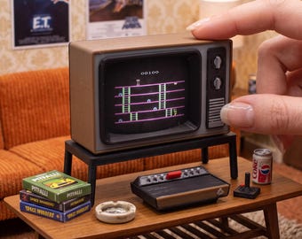 May include: A 1:12 scale miniature brown CRT television set displaying a retro video game screen with a score of 00100. The television sits on a black stand, accompanied by a miniature game console, a joystick, and two game boxes.