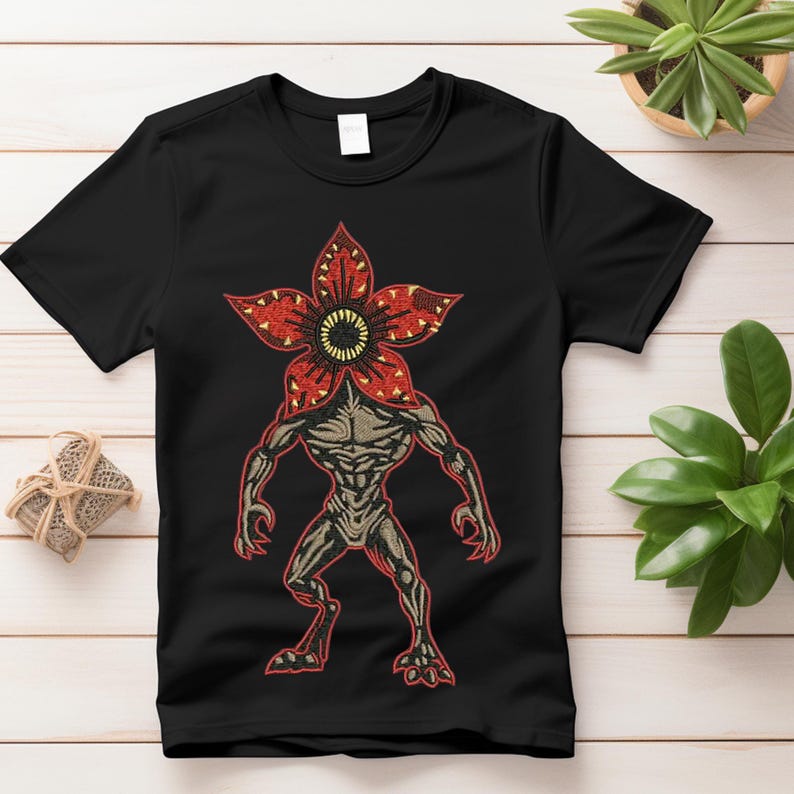 Pode incluir: T-shirt preto com um gr&aacute;fico Demogorgon. O design inclui uma cabe&ccedil;a vermelha em forma de flor com um centro amarelo e um corpo humanoide em tons de castanho e cinzento. A t-shirt &eacute; de gola redonda.
