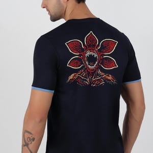 May include: Navy blue t-shirt featuring a Demogorgon graphic on the back. The graphic is red and white with a detailed monster design. The shirt has short sleeves with a light blue trim.