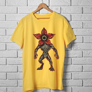 Pode incluir: Camiseta amarela com um gr&aacute;fico Demogorgon. O gr&aacute;fico &eacute; uma ilustra&ccedil;&atilde;o detalhada da criatura com uma cabe&ccedil;a vermelha em forma de flor e um corpo cinza e musculoso. A camiseta est&aacute; pendurada em um cabide de madeira contra uma parede de tijolos brancos.
