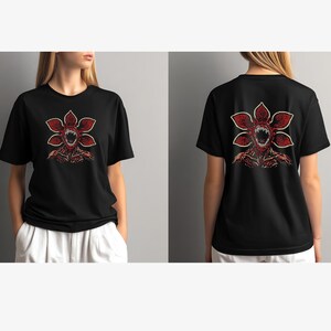 May include: Black t-shirt featuring a red and white Demogorgon graphic on the front and back. The design depicts a flower-like creature with sharp teeth and a menacing expression. The shirt is a short-sleeved crew neck.