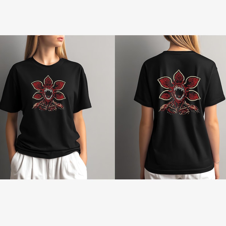 May include: Black t-shirt featuring a red and white Demogorgon graphic on the front and back. The design depicts a flower-like creature with sharp teeth and a menacing expression. The shirt is a short-sleeved crew neck.