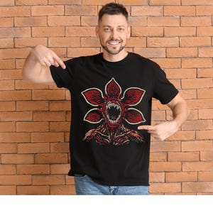 May include: Black t-shirt featuring a Demogorgon graphic. The design showcases a red, flower-like creature with a gaping mouth and sharp teeth. The graphic is centered on the shirt, with a detailed illustration of the creature's head and body.