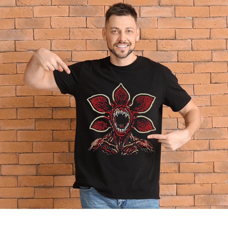 May include: Black t-shirt featuring a Demogorgon graphic. The design showcases a red, flower-like creature with a gaping mouth and sharp teeth. The graphic is centered on the shirt, with a detailed illustration of the creature's head and body.