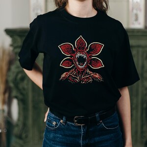 May include: Black t-shirt featuring a Demogorgon graphic. The design showcases a red, flower-like creature with sharp teeth and a detailed, textured body. The graphic is centered on the shirt, offering a bold and eye-catching design.