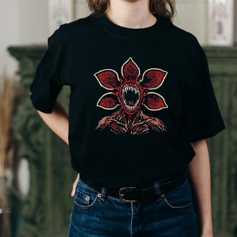 May include: Black t-shirt featuring a Demogorgon graphic. The design showcases a red, flower-like creature with sharp teeth and a detailed, textured body. The graphic is centered on the shirt, offering a bold and eye-catching design.