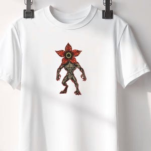 Pode incluir: T-shirt branco com um gr&aacute;fico Demogorgon. O gr&aacute;fico mostra uma criatura com uma cabe&ccedil;a vermelha em forma de flor e um corpo humanoide musculoso em tons de castanho e cinzento. O desenho est&aacute; centrado na t-shirt.