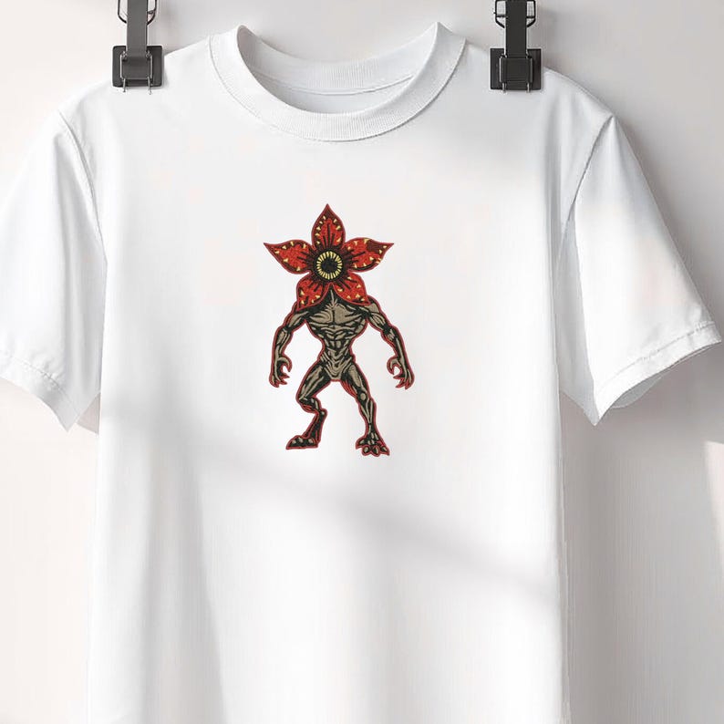 Pode incluir: T-shirt branco com um gr&aacute;fico Demogorgon. O gr&aacute;fico mostra uma criatura com uma cabe&ccedil;a vermelha em forma de flor e um corpo humanoide musculoso em tons de castanho e cinzento. O desenho est&aacute; centrado na t-shirt.