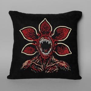 May include: Black square pillow featuring a red and white Demogorgon design. The creature has a flower-like head with sharp teeth and a detailed, textured body. The pillow is a decorative item.