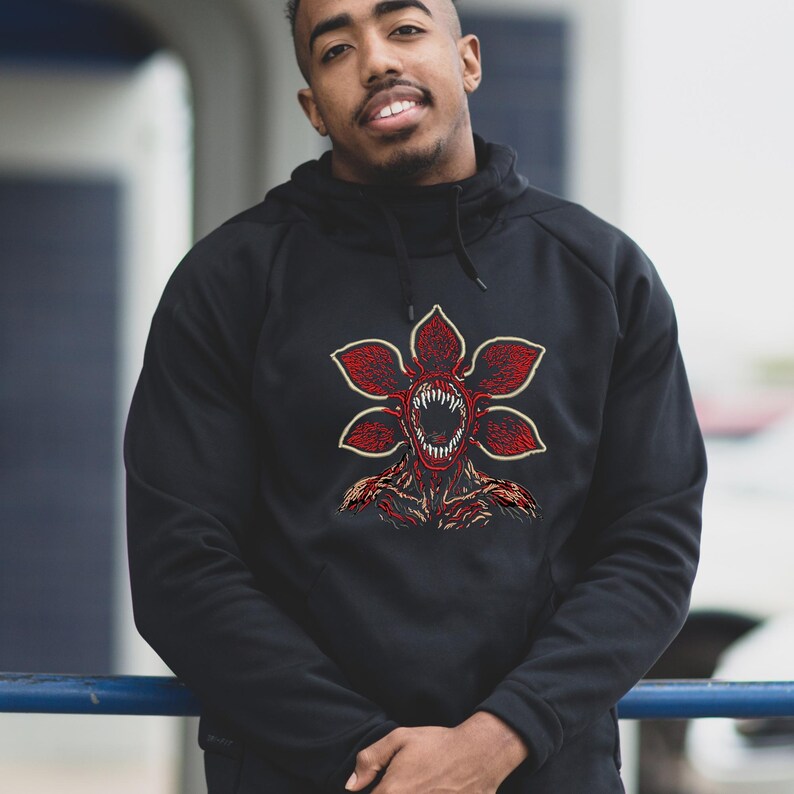 May include: Black hooded sweatshirt featuring a red and white Demogorgon graphic. The graphic is a detailed illustration of the creature's head with open jaws and sharp teeth. The sweatshirt has a drawstring hood and long sleeves.