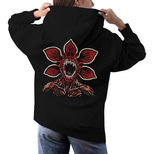 May include: Black hoodie featuring a Demogorgon graphic on the back. The Demogorgon design is red and cream, with a detailed illustration of the creature's face and body. The hoodie has a hood and long sleeves.