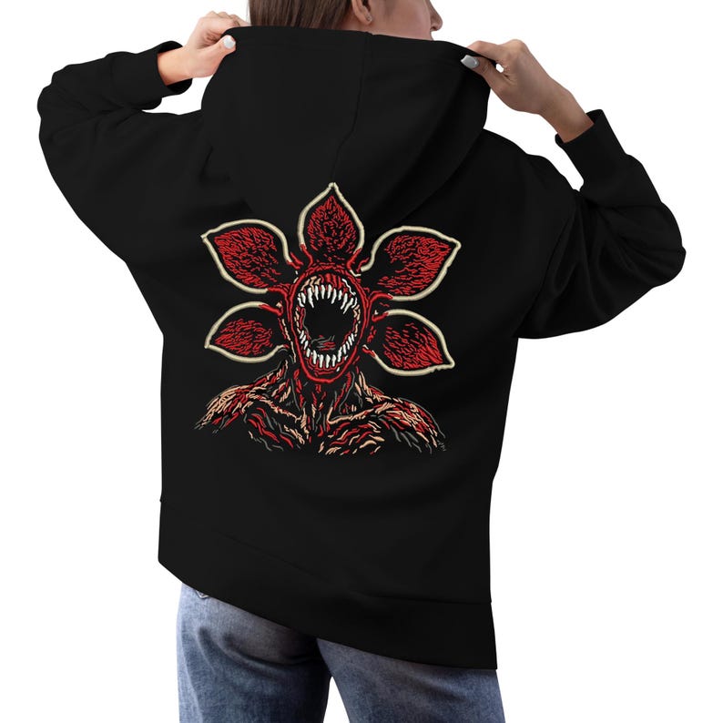 May include: Black hoodie featuring a Demogorgon graphic on the back. The Demogorgon design is red and cream, with a detailed illustration of the creature's face and body. The hoodie has a hood and long sleeves.