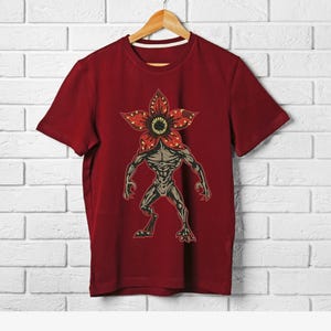 Pode incluir: Uma camiseta bord&ocirc; com um gr&aacute;fico de Demogorgon. O design inclui uma cabe&ccedil;a em forma de flor com p&eacute;talas vermelhas e um centro amarelo, e um corpo humanoide. A camiseta est&aacute; pendurada em um cabide de madeira contra uma parede de tijolos brancos.