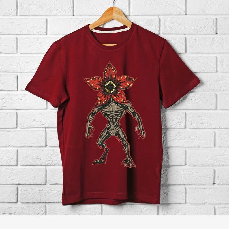 Pode incluir: Uma camiseta bord&ocirc; com um gr&aacute;fico de Demogorgon. O design inclui uma cabe&ccedil;a em forma de flor com p&eacute;talas vermelhas e um centro amarelo, e um corpo humanoide. A camiseta est&aacute; pendurada em um cabide de madeira contra uma parede de tijolos brancos.