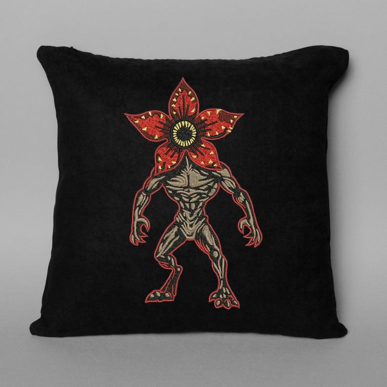 Pode incluir: Almofada quadrada preta com um design bordado de Demogorgon. A criatura tem uma cabe&ccedil;a vermelha em forma de flor com um centro amarelo e um corpo cinza e musculoso. A almofada &eacute; um item decorativo.