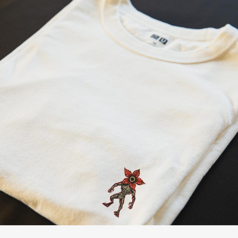 Pode incluir: Camiseta creme com um gr&aacute;fico Demogorgon. O gr&aacute;fico &eacute; uma ilustra&ccedil;&atilde;o detalhada da criatura com uma cabe&ccedil;a vermelha em forma de flor e um corpo cinza. A camiseta est&aacute; dobrada, mostrando a gola redonda e o gr&aacute;fico na frente.