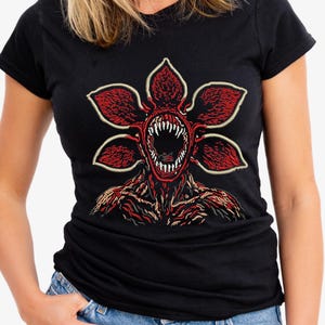 May include: Black t-shirt featuring a red and white graphic of a Demogorgon from the Stranger Things series. The Demogorgon has a flower-like head with sharp teeth and a detailed, textured body. The design is centered on the shirt.