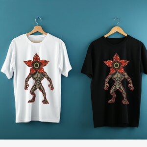 Pode incluir: Duas camisetas, uma branca e outra preta, cada uma com um gr&aacute;fico Demogorgon. O gr&aacute;fico retrata uma figura humanoide com uma cabe&ccedil;a em forma de flor em tons de vermelho, marrom e bege. As camisetas s&atilde;o exibidas em cabides de madeira.