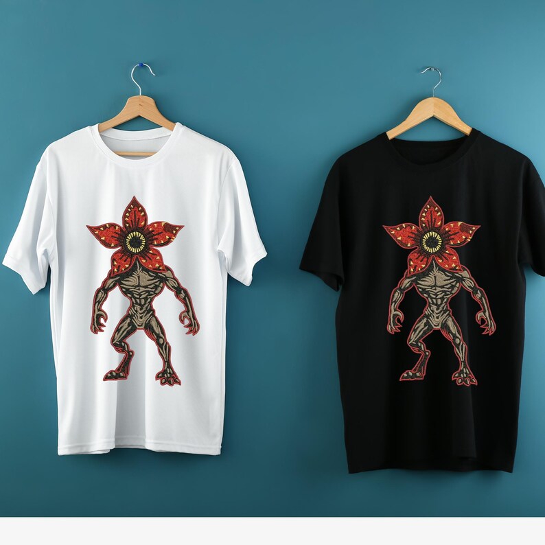 Pode incluir: Duas camisetas, uma branca e outra preta, cada uma com um gr&aacute;fico Demogorgon. O gr&aacute;fico retrata uma figura humanoide com uma cabe&ccedil;a em forma de flor em tons de vermelho, marrom e bege. As camisetas s&atilde;o exibidas em cabides de madeira.