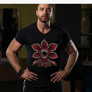 May include: Black V-neck t-shirt featuring a red and white Demogorgon graphic. The graphic depicts a monster with a flower-like head and sharp teeth. The shirt is made of a soft material and is suitable for casual wear.