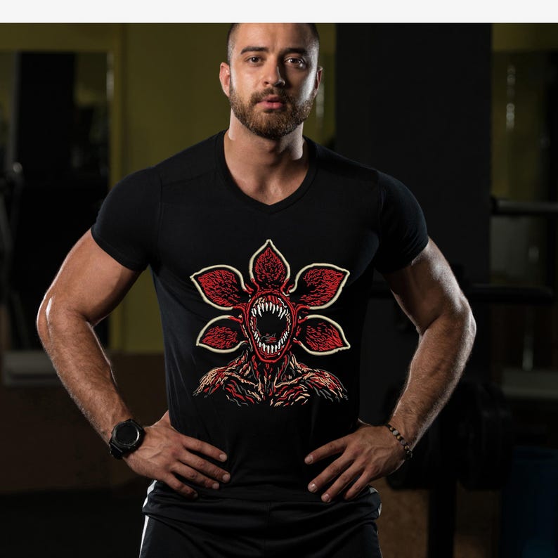 May include: Black V-neck t-shirt featuring a red and white Demogorgon graphic. The graphic depicts a monster with a flower-like head and sharp teeth. The shirt is made of a soft material and is suitable for casual wear.