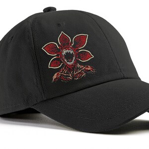 May include: Black baseball cap featuring a red and white Demogorgon flower design. The embroidered graphic depicts a monster with a toothed mouth and petal-like shapes. The cap has a curved brim and a button on top.
