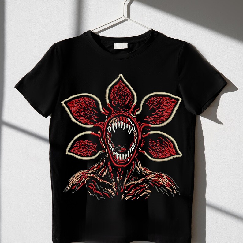 May include: Black t-shirt featuring a Demogorgon graphic. The design showcases a red, flower-like head with sharp teeth and a detailed body. The graphic is outlined in a cream color.