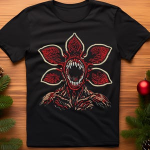 May include: Black t-shirt featuring a red and white graphic of a Demogorgon from the popular science fiction horror series. The design shows the creature's open mouth with sharp teeth and a flower-like head.