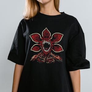 May include: Black t-shirt featuring a red and white graphic of a Demogorgon from the popular science fiction series. The graphic depicts the creature's flower-like head with sharp teeth and a detailed body. The shirt is a casual, oversized fit.