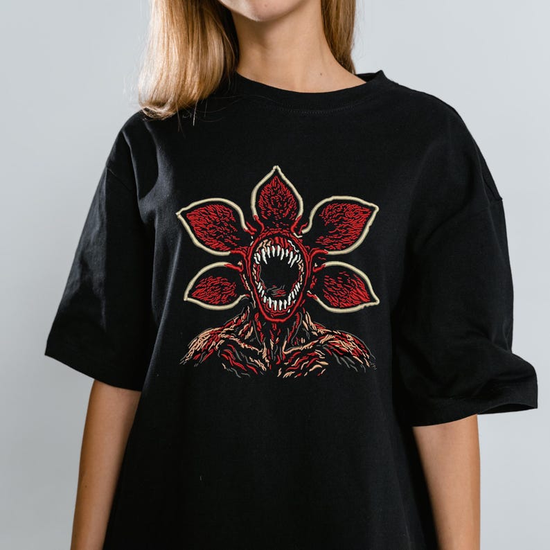 May include: Black t-shirt featuring a red and white graphic of a Demogorgon from the popular science fiction series. The graphic depicts the creature's flower-like head with sharp teeth and a detailed body. The shirt is a casual, oversized fit.