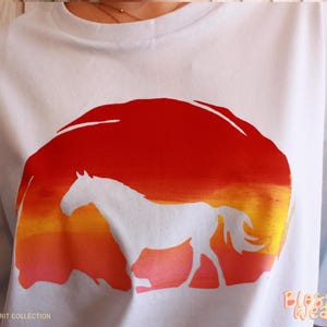 Może przedstawiać: Biała koszulka z sylwetką konia na tle gradientu zachodu słońca w kolorach czerwonym, pomarańczowym i żółtym. Tekst "HORSE SPIRIT COLLECTION" znajduje się w lewym dolnym rogu. Tekst "Blossom Weave" znajduje się w prawym dolnym rogu.