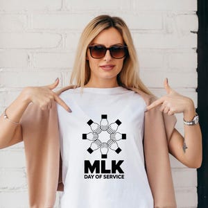 May include: White t-shirt with a black graphic of hands forming a circle, with the text "MLK DAY OF SERVICE" below. The shirt is made of a soft material.