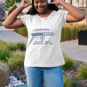 Op de afbeelding: Wit V-hals T-shirt met een blauw en witte afbeelding met de tekst "LOADING.." met een voortgangsbalk en de tekst "PLEASE WAIT CONNECTED AT 56K". Het shirt wordt gecombineerd met een blauwe jeans.
