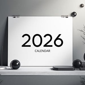 Op de afbeelding: Een witte 2026 kalender met het woord "KALENDER" onder de cijfers. De afbeelding bevat een klok, decoratieve bollen en een vaas met takken. De achtergrond is neutraal grijs.