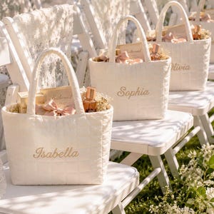 May include: A series of white quilted tote bags with two handles, each featuring an embroidered name on the front. The bags are filled with gift items including small bottles, cards labeled Bridesmaid, and ribbons, resting on white chairs.