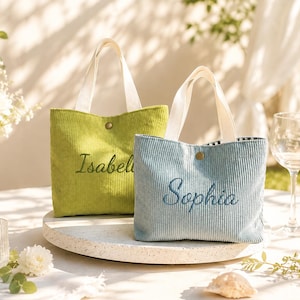 Könnte beinhalten: Zwei Cord-Tragetaschen mit weißen Stoffhenkeln und einem einzelnen messingfarbenen Druckknopfverschluss. Die lindgrüne Tasche ist mit dem Namen Isabelle bestickt, während die hellblaue Tasche den Namen Sophia in Schreibschrift trägt.