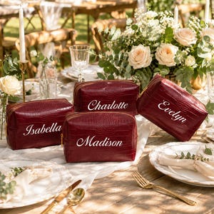 May include: Three deep red, rectangular, faux-croc textured cosmetic bags with gold-tone zippers. Each bag features a white embroidered name: Isabella, Charlotte, Madison, or Evelyn, displayed in a cursive script font on the front surface.
