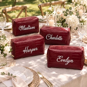 May include: Four rectangular burgundy cosmetic bags with a crocodile-embossed texture and gold-tone zippers. Each bag features a white embroidered name on the front: Madison, Charlotte, Harper, and Evelyn, written in a cursive script font.