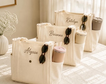 May include: Three off-white canvas tote bags with external pockets, each personalized with a name in dark brown script: Alyssa, Brianna, and Katelyn. Each bag features a pair of dark-rimmed sunglasses and a solid-colored tumbler in the side pocket.