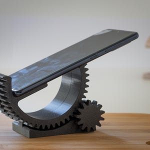 Könnte beinhalten: Ein schwarzes Smartphone liegt auf einem dunkelgrauen, zahnradförmigen Telefonständer. Der Ständer besteht aus 3D-gedrucktem Material und hat ein einzigartiges Design mit ineinandergreifenden Zahnrädern und einer strukturierten Oberfläche. Das Telefon ist abgewinkelt.