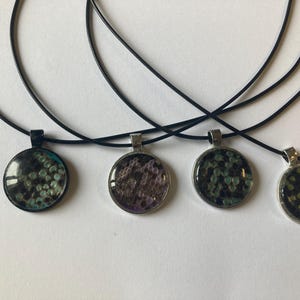 May include: Four round pendant necklaces with black cords. The pendants have silver-coloured frames and feature various abstract designs in shades of green, purple, and black. The necklaces are displayed on a white surface.