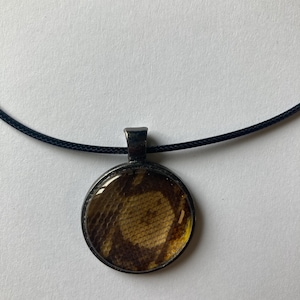 Könnte beinhalten: Eine runde Anhängerhalskette mit schwarzem Band. Der Anhänger hat einen dunklen Metallrahmen und ein kreisförmiges Design mit einem braunen und gelben Muster. Die Halskette wird auf einer weißen Oberfläche präsentiert.