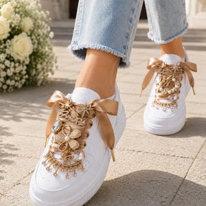 May include: A pair of white sneakers featuring wide, metallic gold satin ribbon laces. The laces are adorned with decorative gold-toned charms, including a flower shape, star accents, and dangling clear crystals that drape over the front.