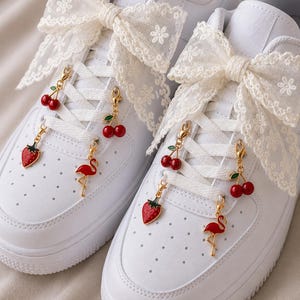 May include: White sneakers adorned with lace bows and fruit-themed charms. The charms include red cherries, strawberries, and a flamingo. The sneakers have white laces and a white sole.