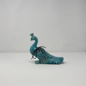 May include: A turquoise ceramic peacock figurine with a detailed tail and a decorative crest. The peacock has a long, curved neck and is adorned with a dark brown string. The figurine is a decorative item.