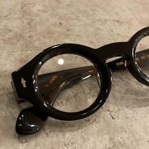 Puede incluir: Gafas de carey marrón oscuro con monturas redondas. La patilla presenta un pequeño emblema de flecha plateada en el lateral. El interior de la patilla tiene impreso el texto 23106 y C02 CE. Las lentes son transparentes y la montura es brillante.