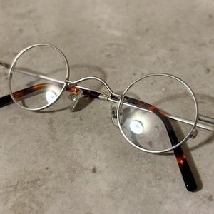May include: Round spectacles featuring thin silver metal frames and clear lenses. The temples are finished with dark brown and black tortoiseshell-patterned acetate tips. The bridge is curved, and the nose pads are made of clear plastic.