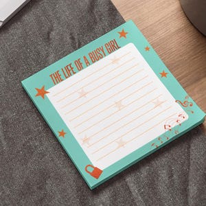 以下が含まれることがあります： 「THE LIFE OF A BUSY GIRL」の文字が入った、水色の縁取りの正方形のメモ帳。星のアクセントと小さな赤い南京錠のアイコンが付いた罫線入りのページがあります。隅には音符のグラフィックがあります。