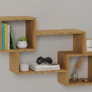 May include: A modern, geometric wooden wall shelf with multiple compartments. The shelf holds books, a small potted plant, a candle, a stack of books, a decorative object, a framed photo, and a vase with dried flowers. The shelf is a light brown color.