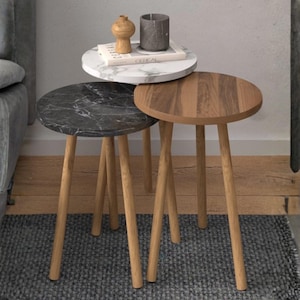May include: Three nesting side tables with round tops. One table has a white marble top, another has a black marble top, and the third has a wood grain top. All three tables have light-colored wooden legs. Decorative items sit on the top table.